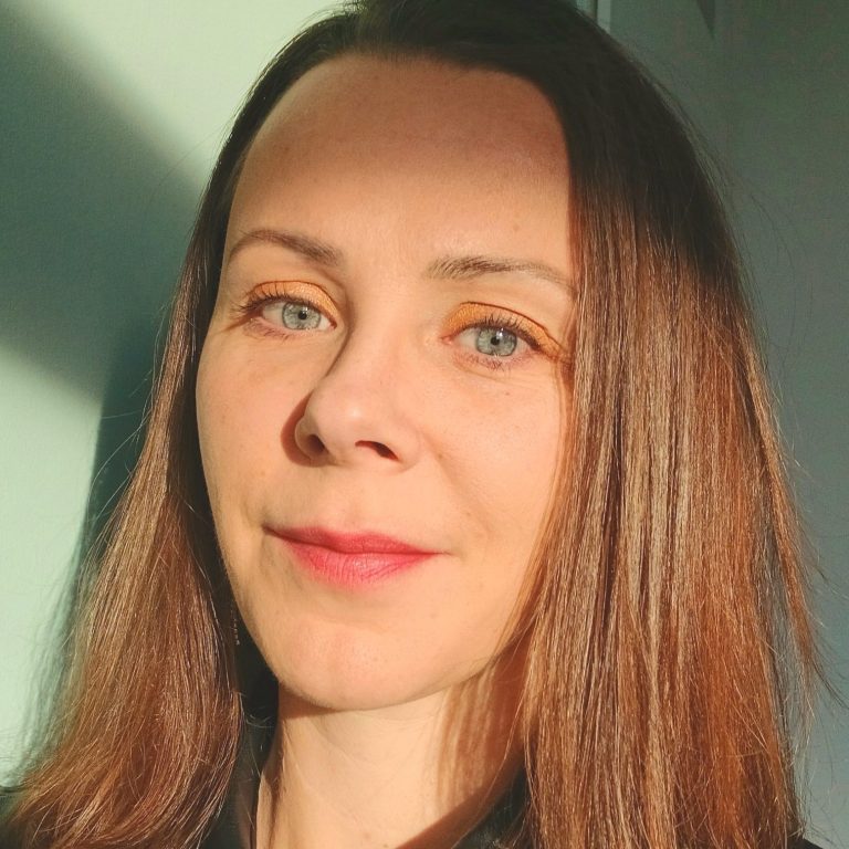 Portret van een vrouw met lang, bruin haar en een lichte huid, tegen een lichte achtergrond.